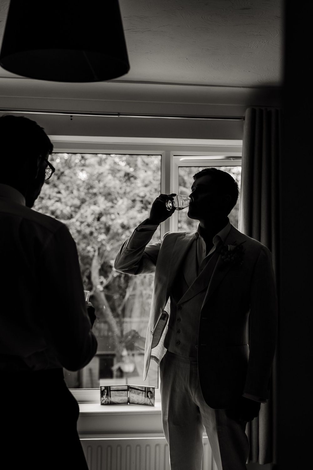Black and white photo of two silhouettes at a window, with one person taking a drink.
