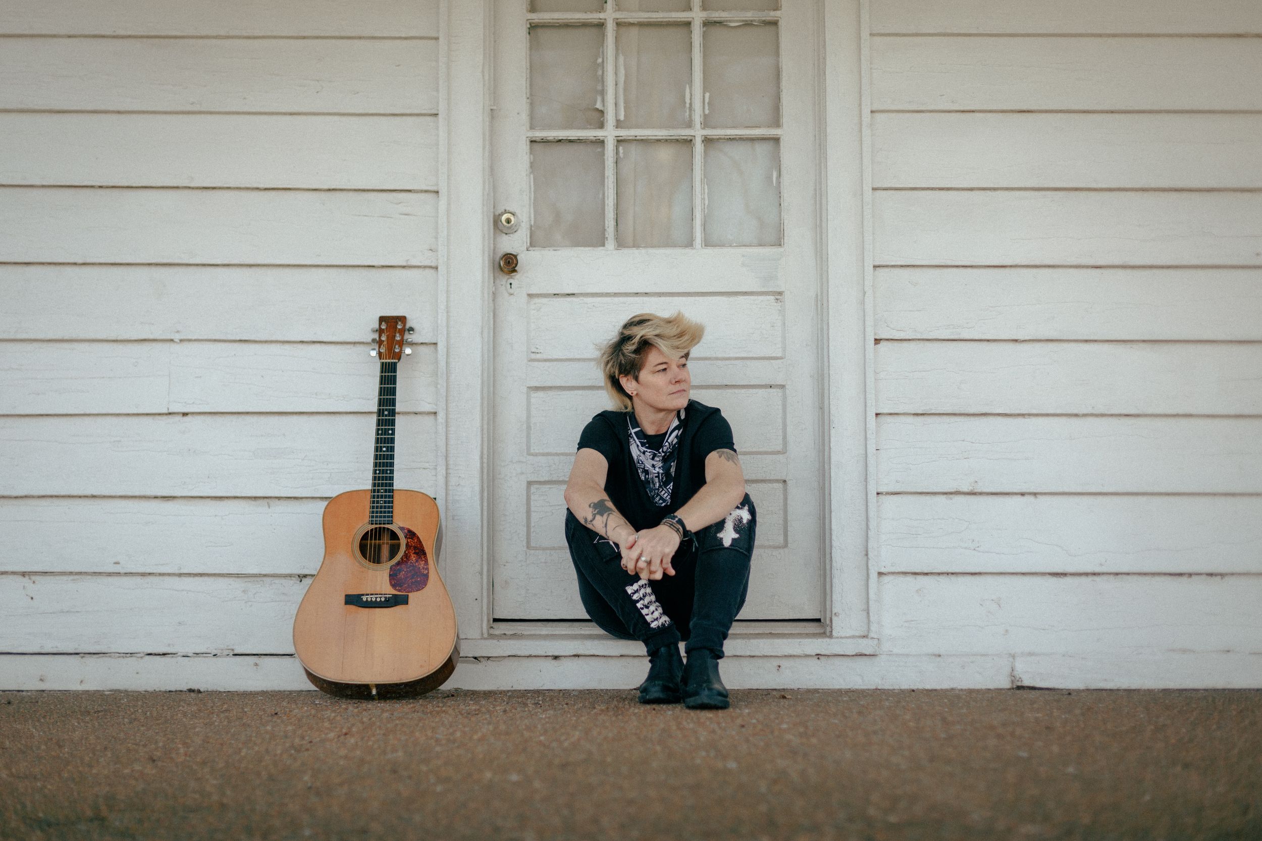 Acoustic guitar leans against white wooden door frame while musician Sonia Leigh sits on porch step.