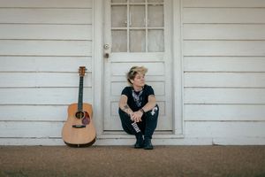 Acoustic guitar leans against white wooden door frame while musician Sonia Leigh sits on porch step.