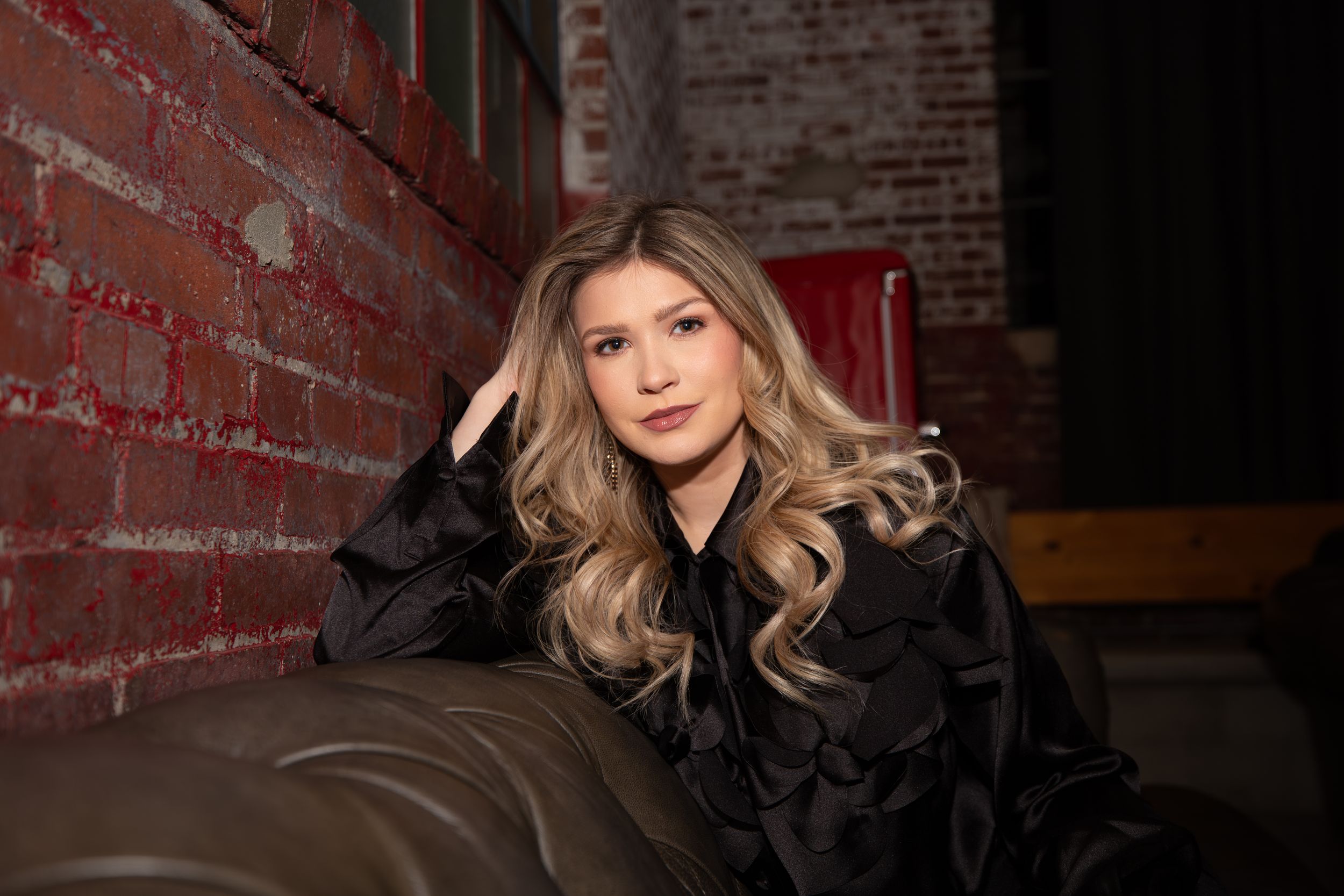 Country artist Charity Bowden lounges on a brown leather couch against a red brick wall in a dark indoor setting.
