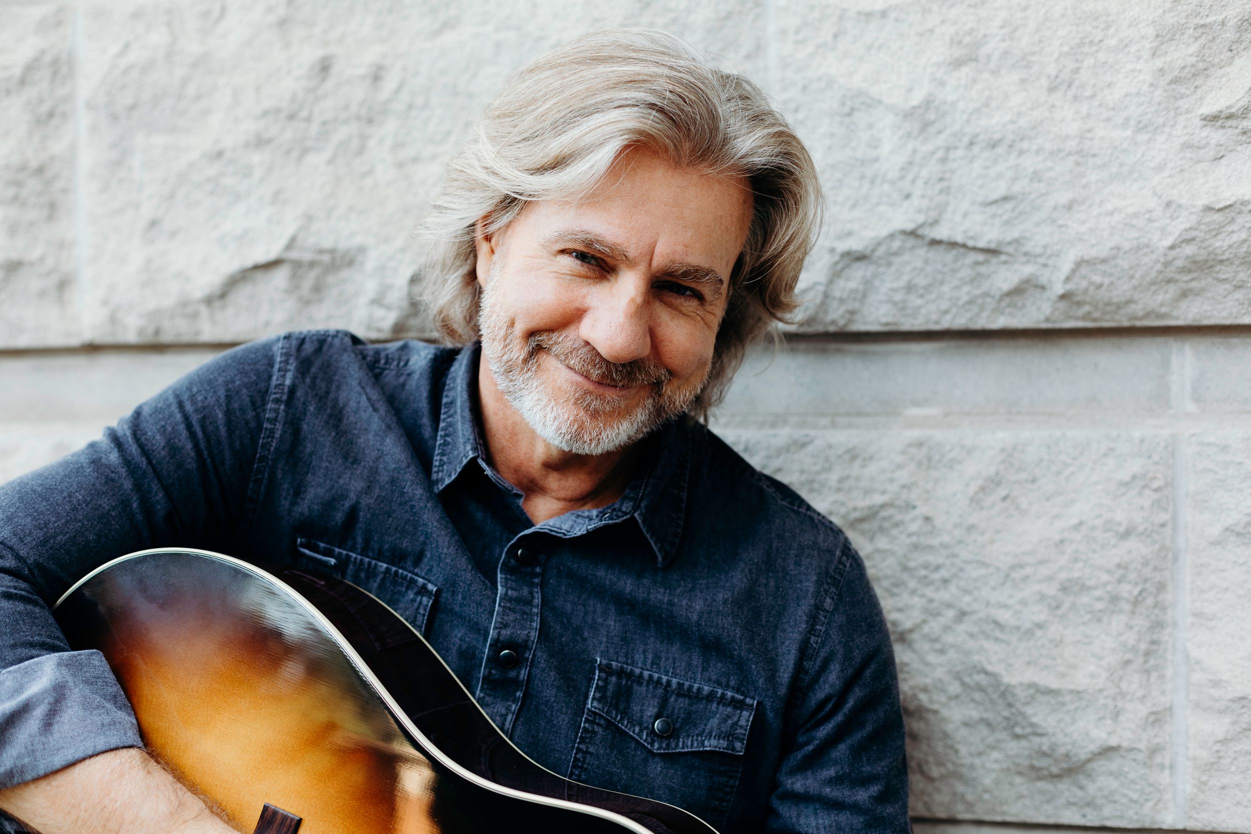 Smiling musician Dwayne O'Brien of Little Texas wearing denim shirt holds acoustic guitar against white textured wall.