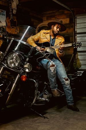 Musician Michael B Whit in a yellow jacket playing guitar while seated on a motorcycle in a rustic garage setting.