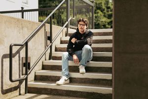 A person sits casually on concrete steps wearing jeans, sneakers and a dark sweater on a sunny day.