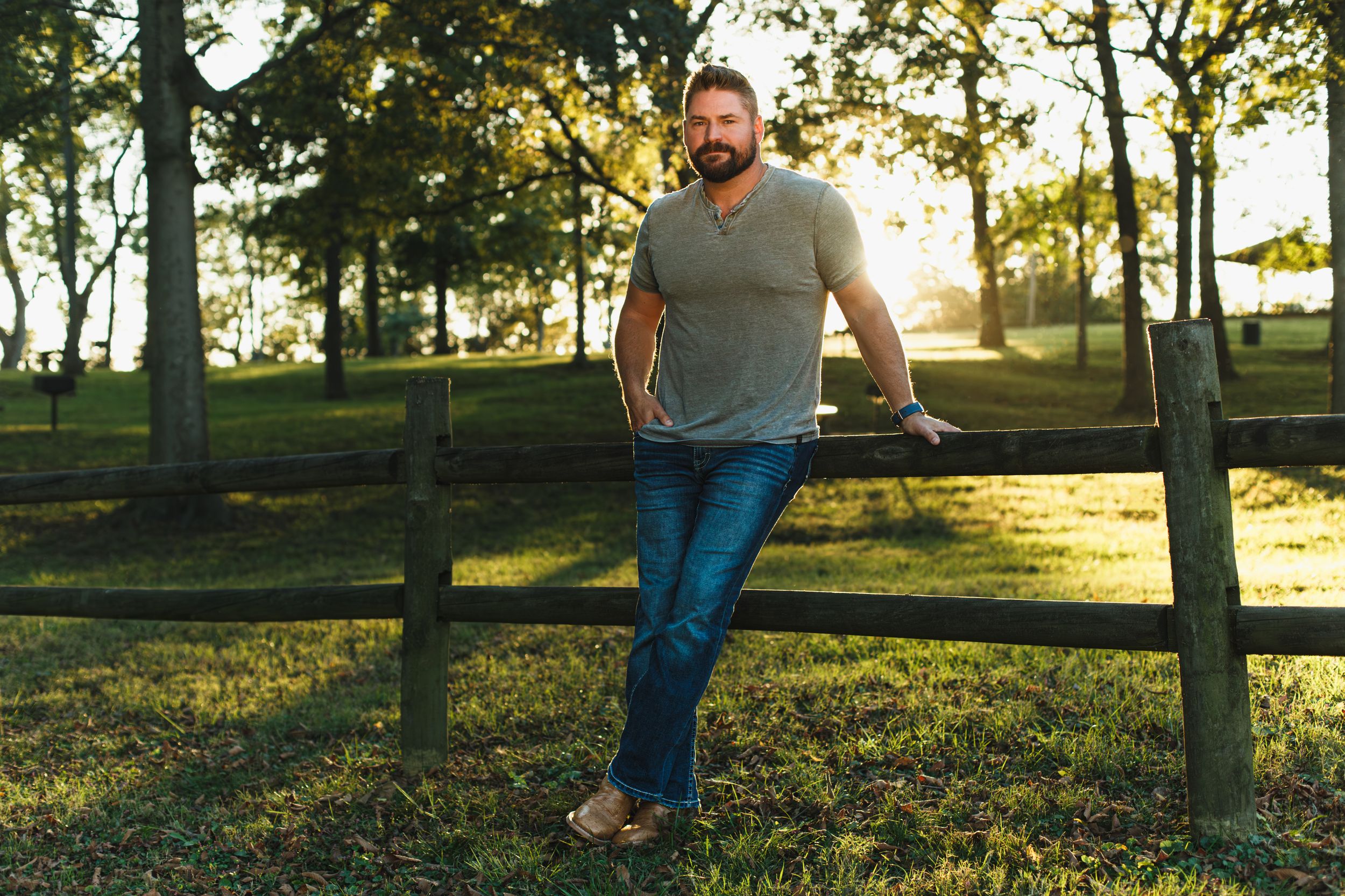 Country artist Jimmy Charles leans against a black fence in a park at sunset, wearing jeans and a grey t-shirt in warm golden light.