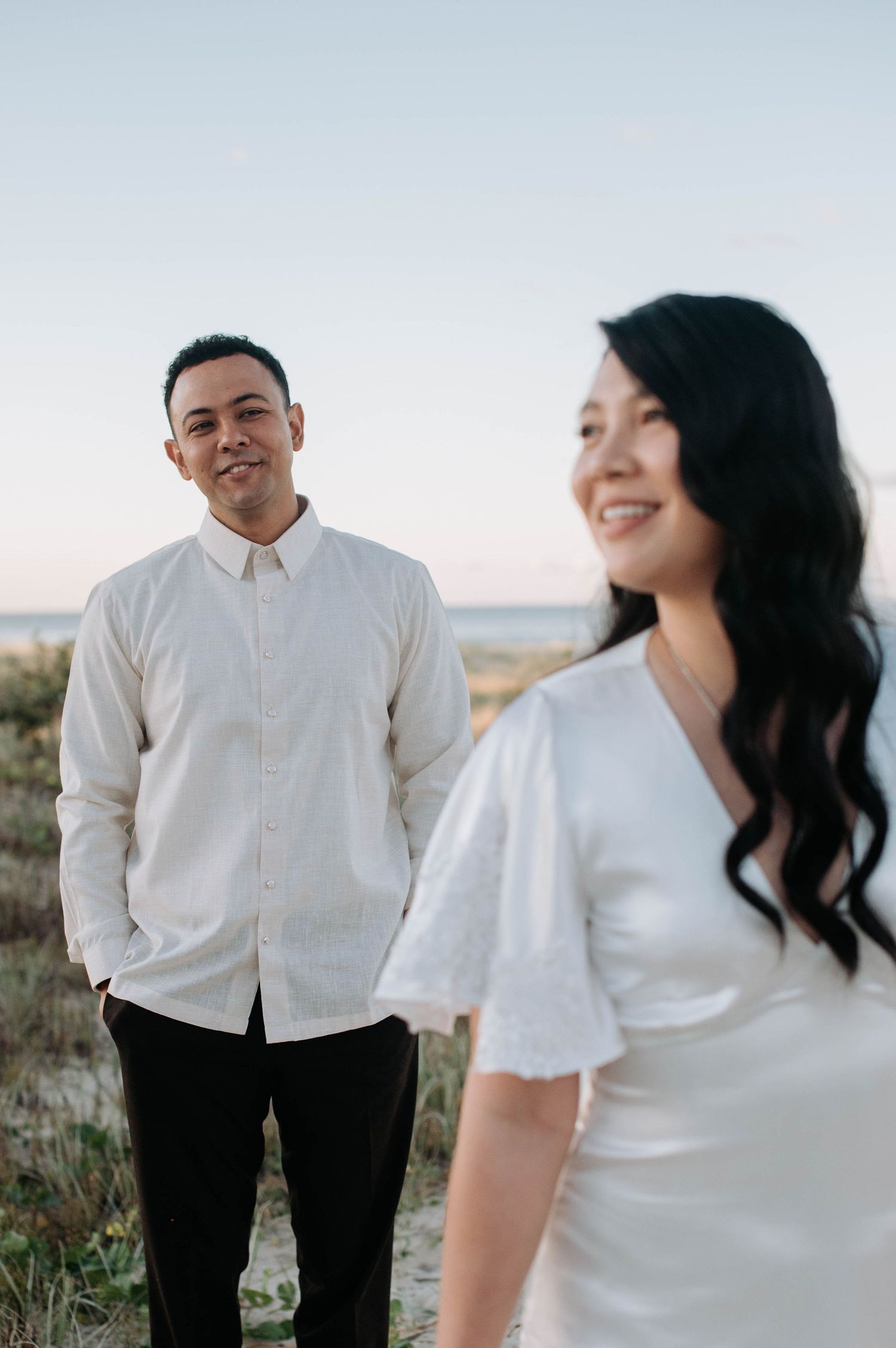 A couple in white outfits pose together outdoors during a sunset engagement photoshoot in a natural setting.