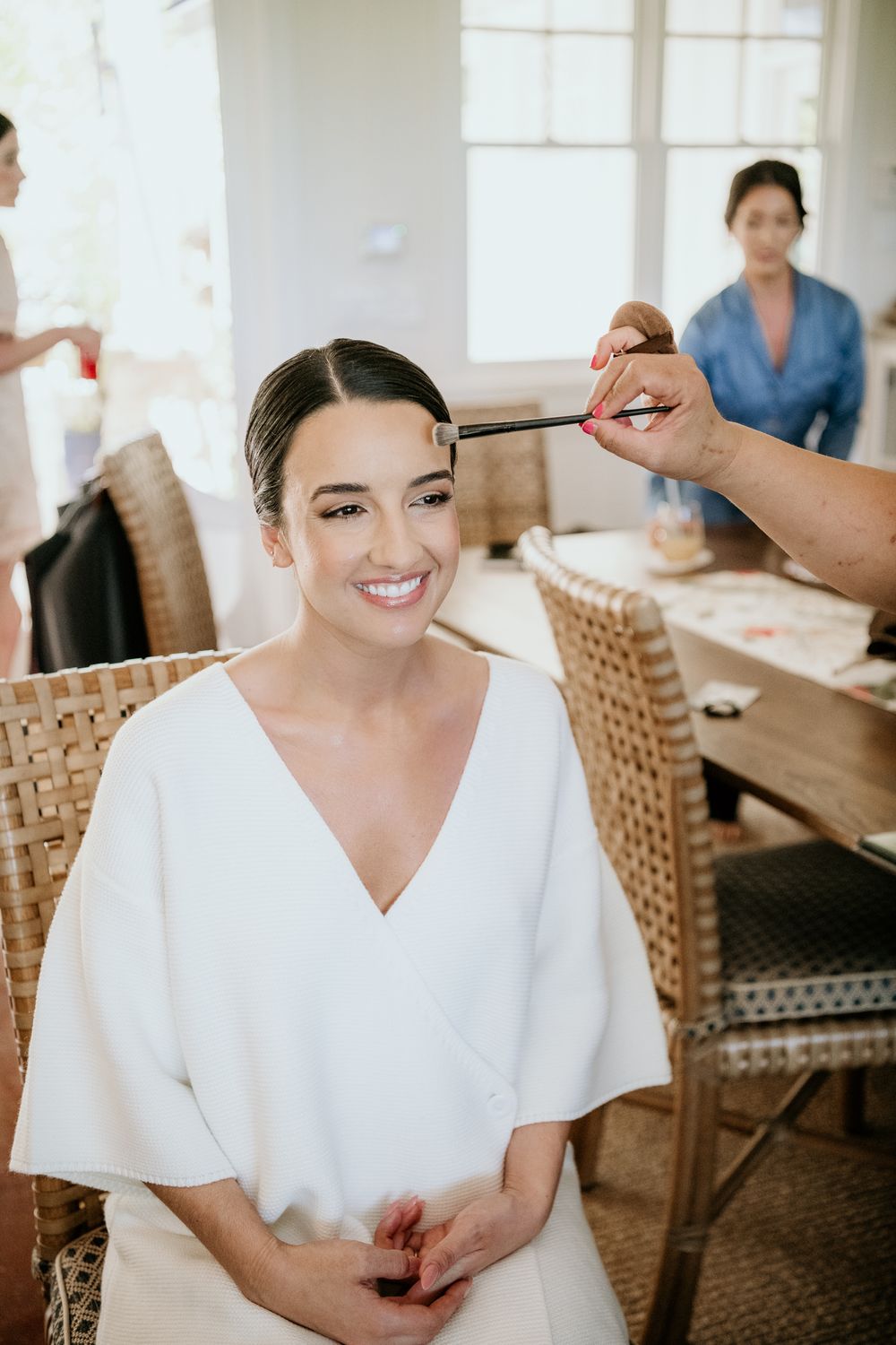 Someone in a white v-neck dress smiles while getting their hair and makeup done in a room with wicker chairs.