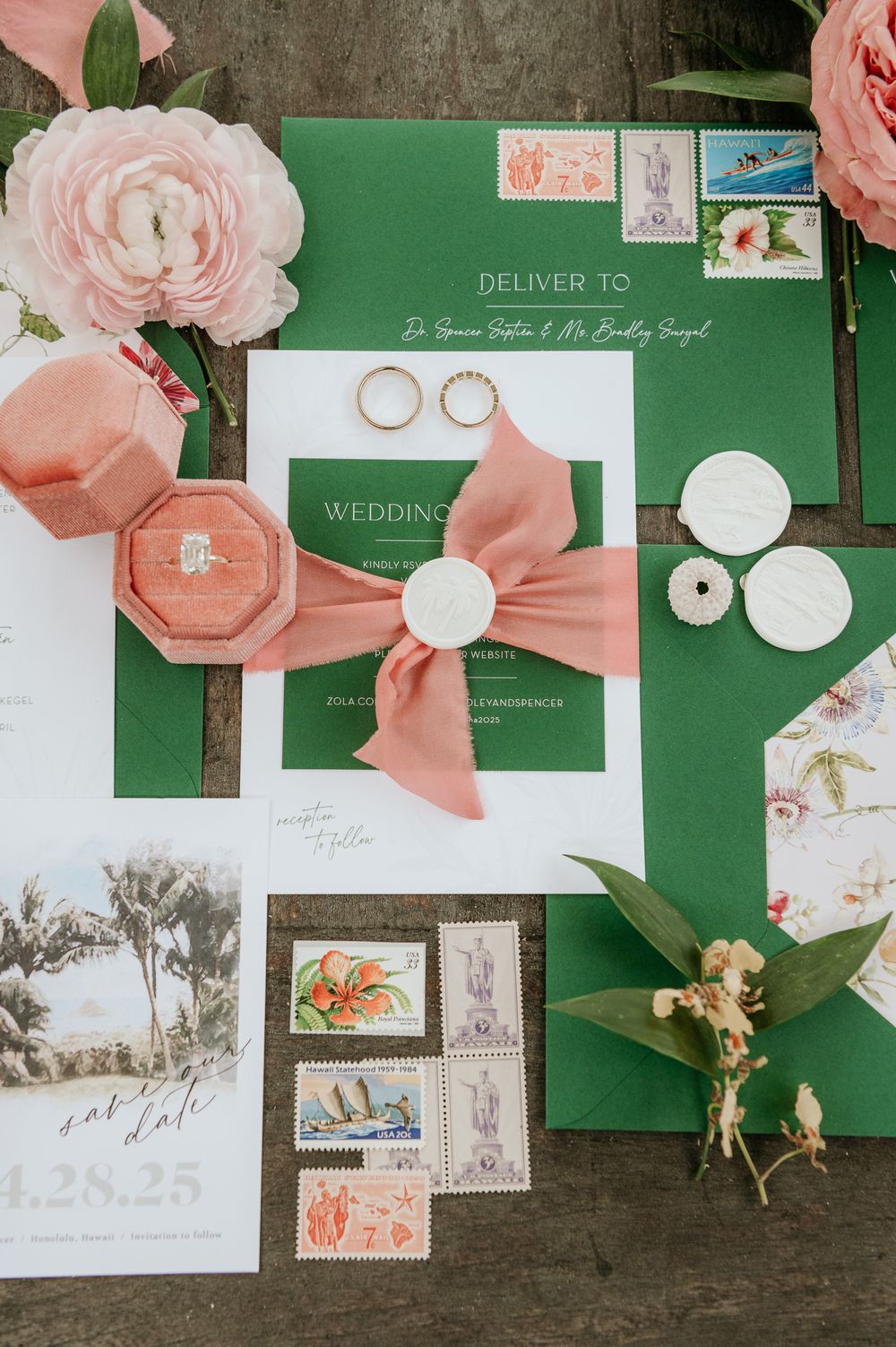 Wedding stationery flatlay with green envelopes, peach ribbons, stamps, and pink roses on dark wooden surface.