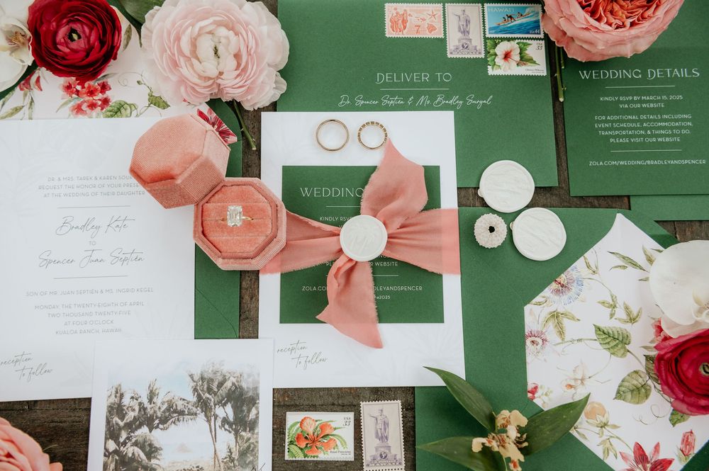Wedding invitation suite with pink and red roses, gold rings, and emerald green stationery displayed on a flat surface.