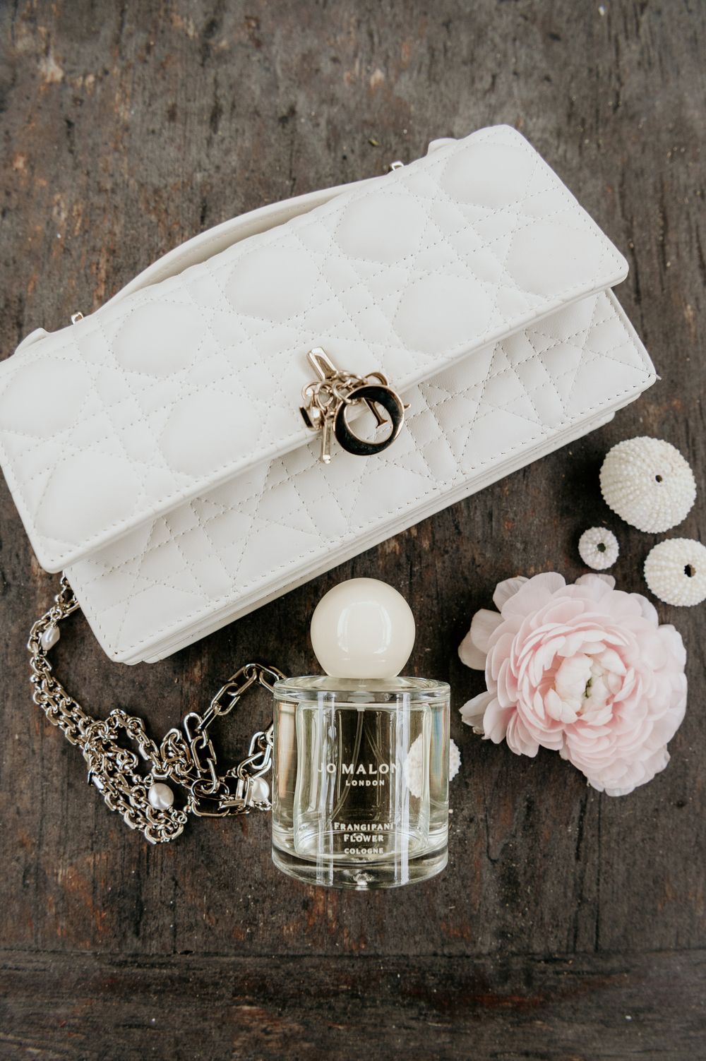 White quilted handbag with chain strap arranged with perfume bottle and pink rose on rustic wooden surface.