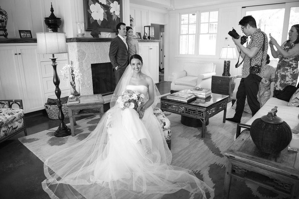 Black and white photo showing bride in flowing white gown being photographed in vintage-style interior room setting.