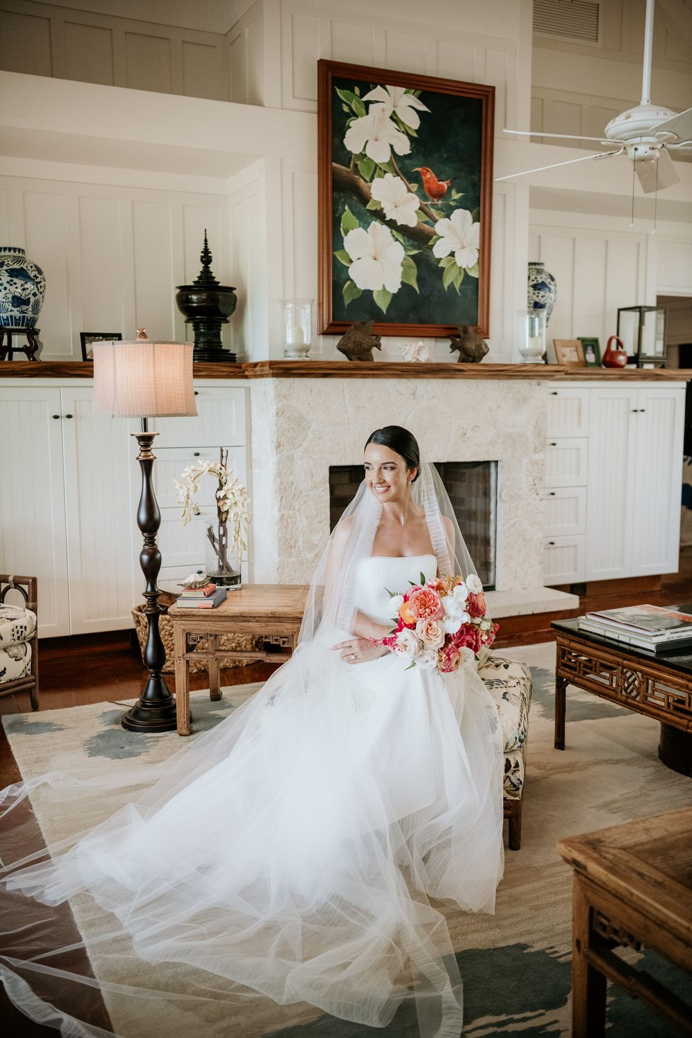 Elegant bride in flowing white gown and veil seated in vintage-style room with floral artwork and warm lighting.