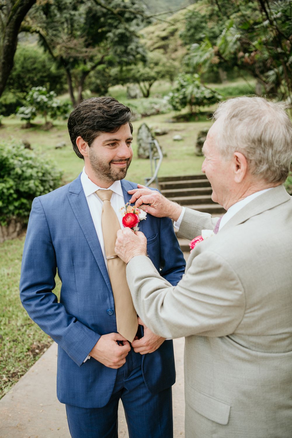 An older gentleman in beige suit helps adjust a pink boutonniere on a younger man wearing a navy blue suit outdoors.