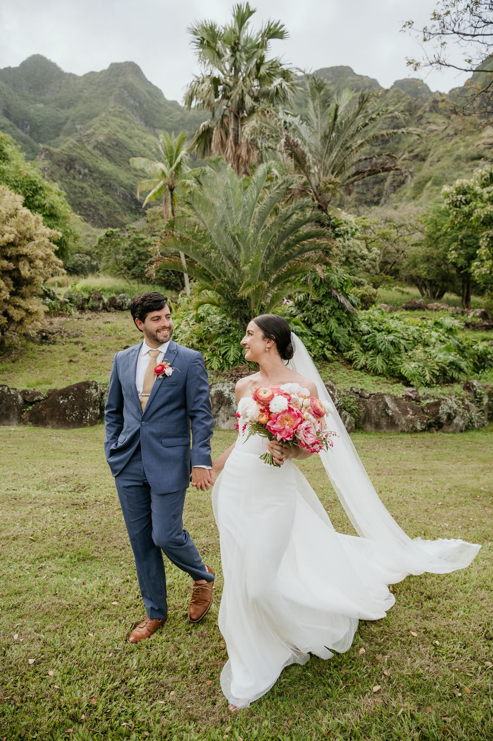 A wedding couple walks on grass with lush tropical mountains and palm trees in the background at Kualoa Ranch Hawaii.