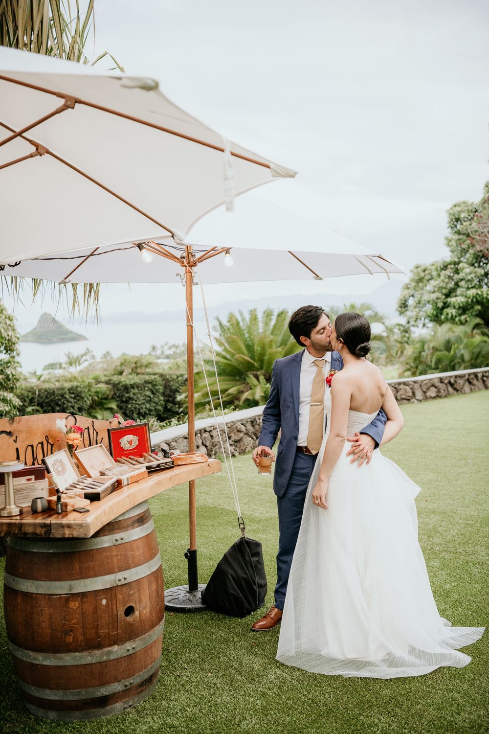 A couple celebrates at an outdoor wedding reception with white umbrellas, wine barrels and tropical greenery decor.