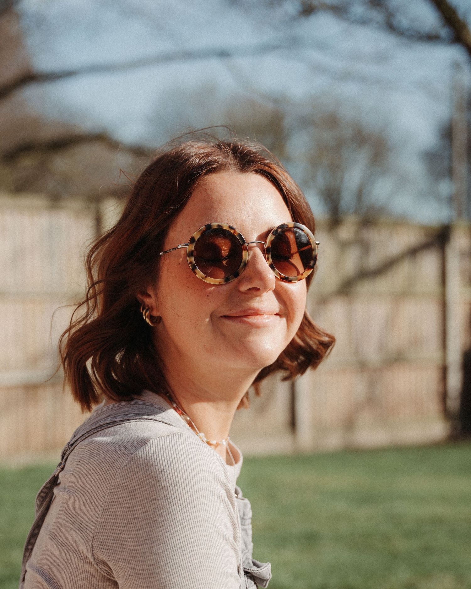 Person in round tortoiseshell sunglasses smiling outdoors on a sunny day.