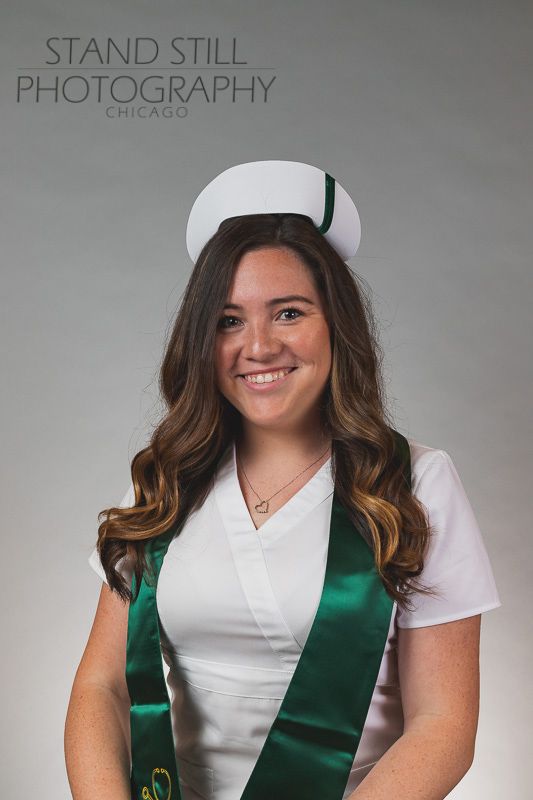 Graduate wearing white uniform with green sash and nurse's cap in studio portrait.