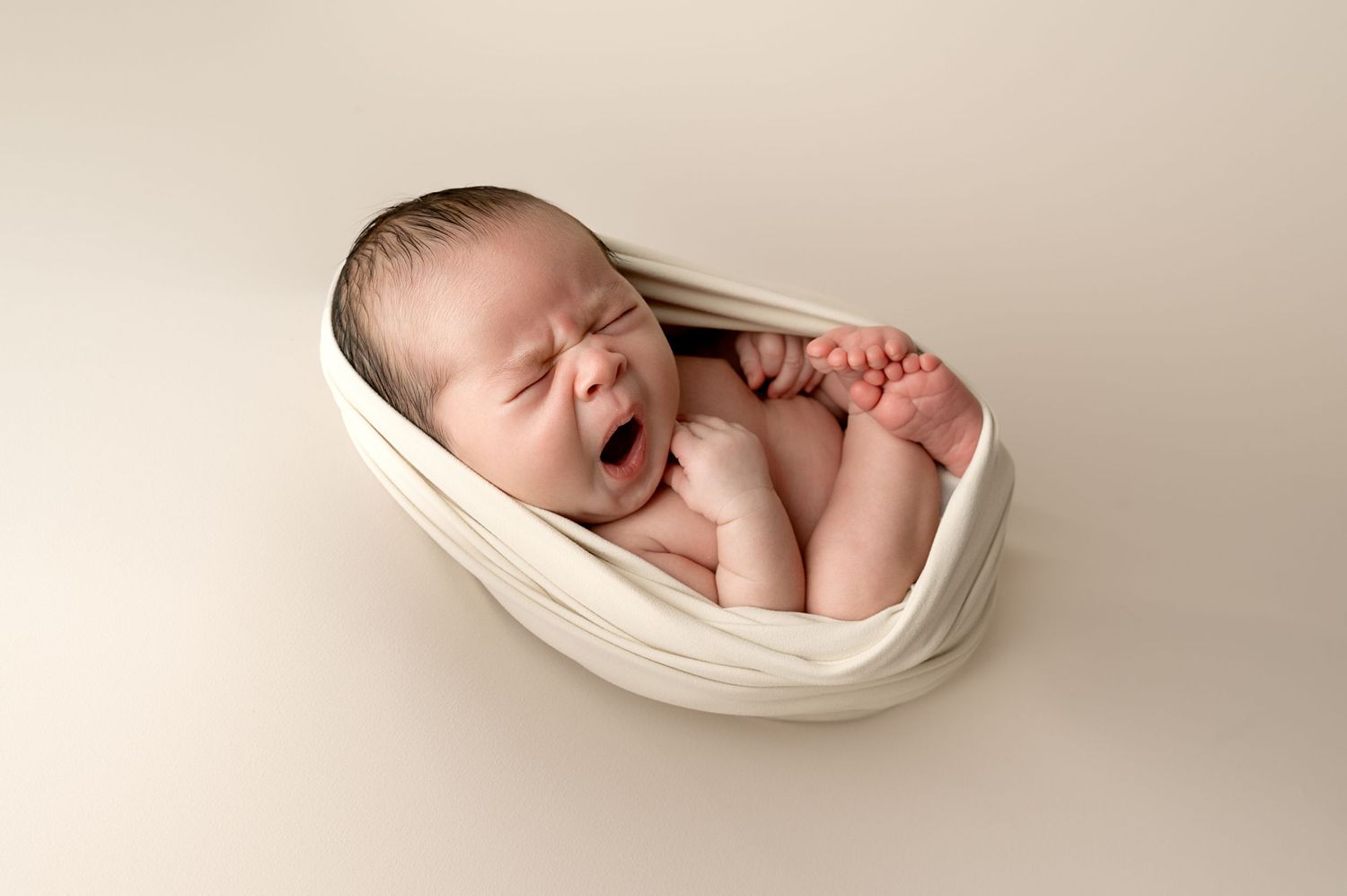 Yawning newborn baby wrapped in white fabric lying in cream-colored nest prop.
