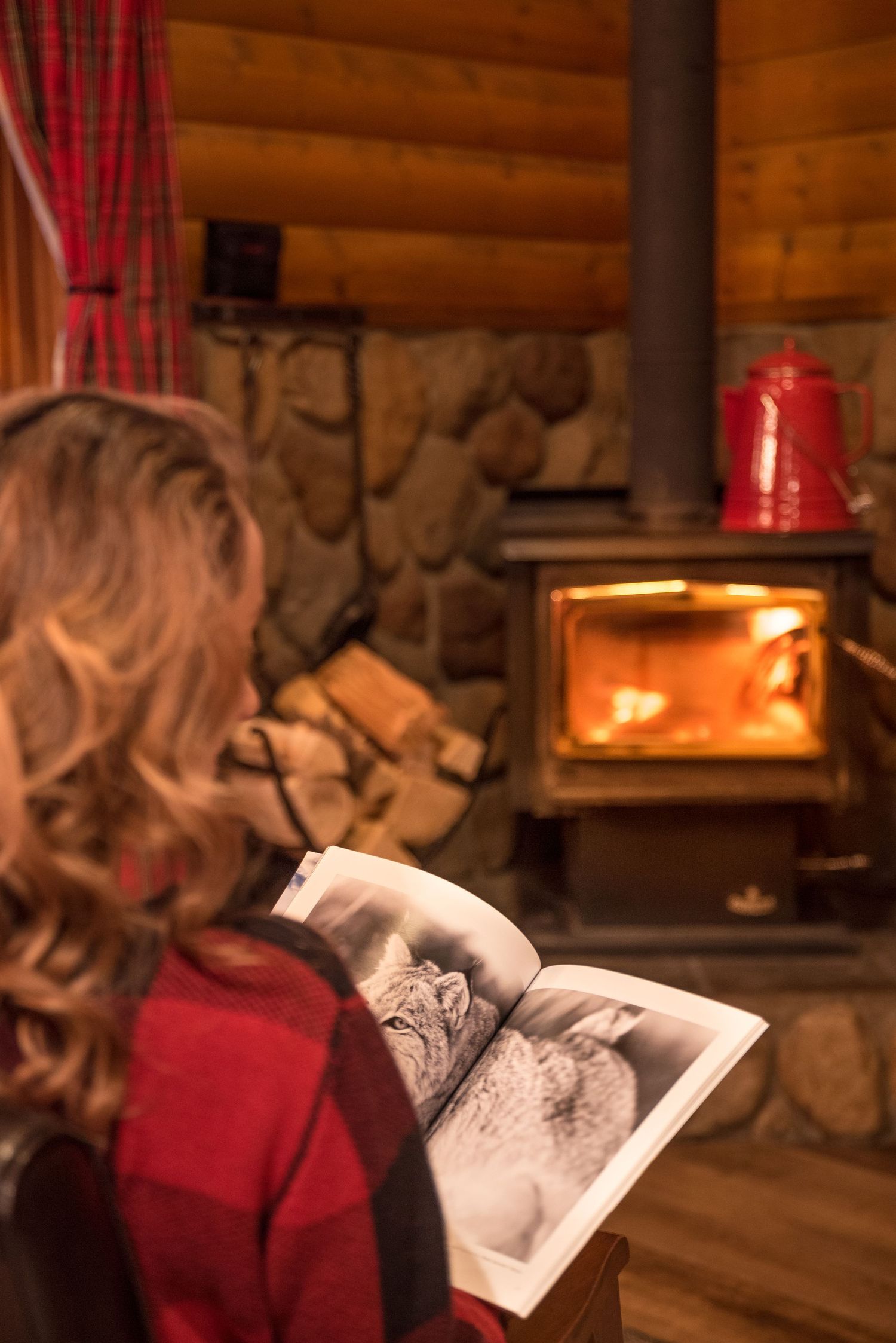 Someone reading a book by a cozy stone fireplace with burning logs in a rustic cabin interior.