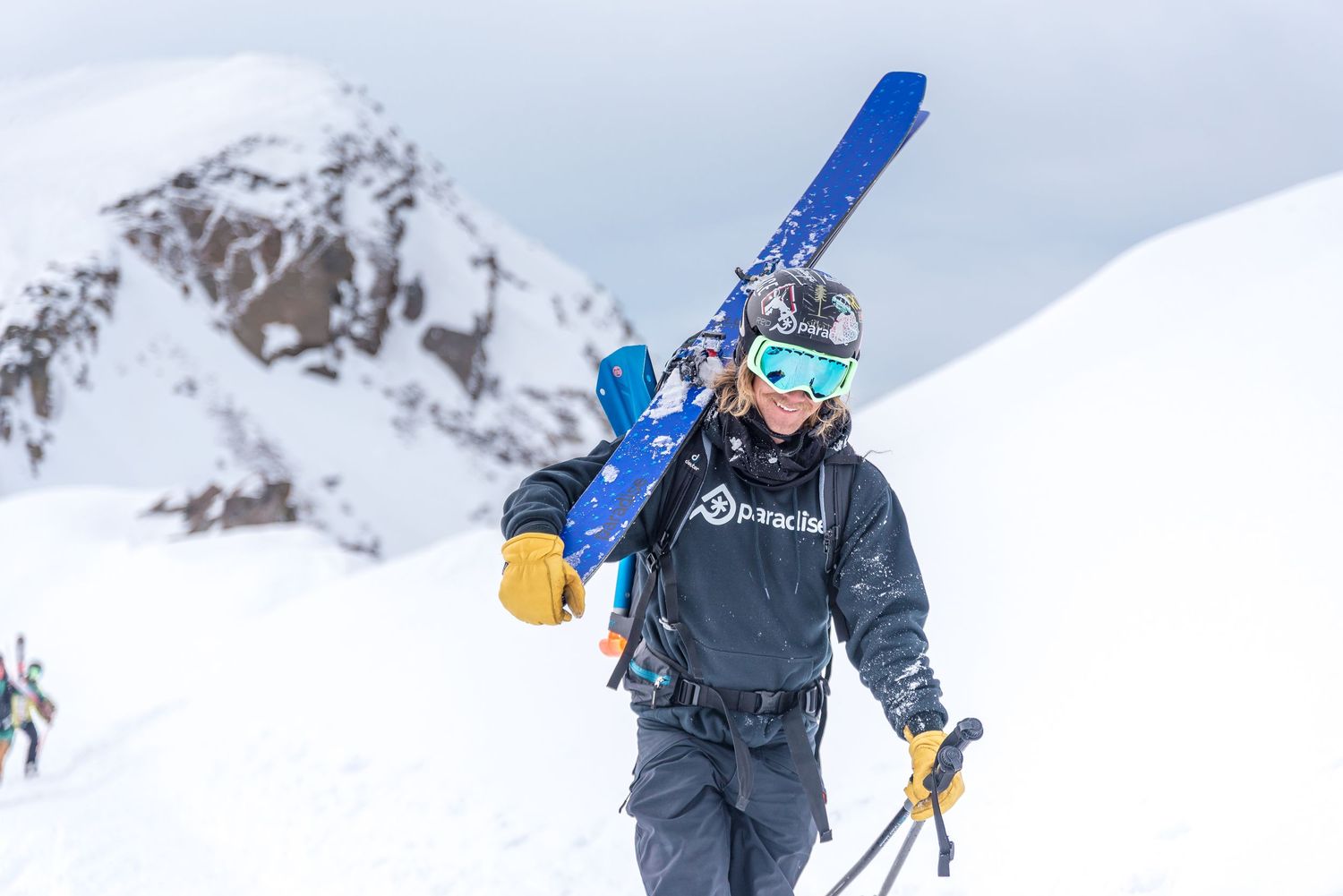 Skier carrying blue skis walks up snowy mountain slope in stormy winter conditions wearing black jacket and goggles.