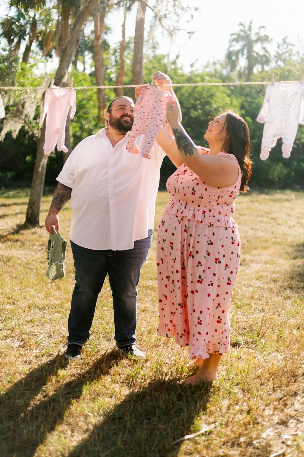 A couple hangs pink baby clothes on a clothesline outdoors in a sunlit backyard with palm trees.