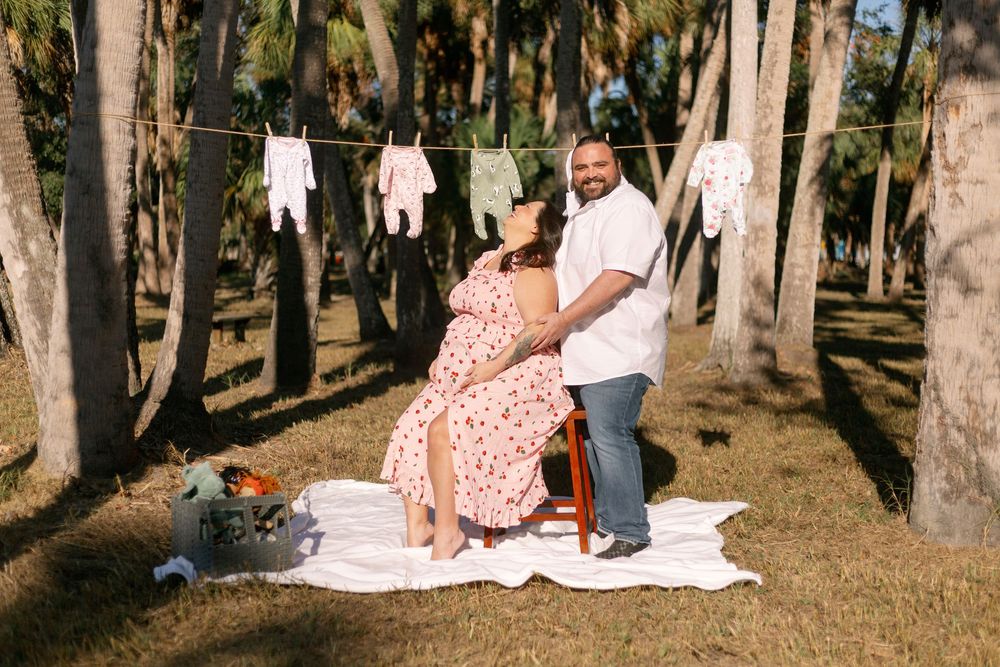A couple poses for a pregnancy photo shoot in a wooded area with baby clothes hanging on a clothesline.
