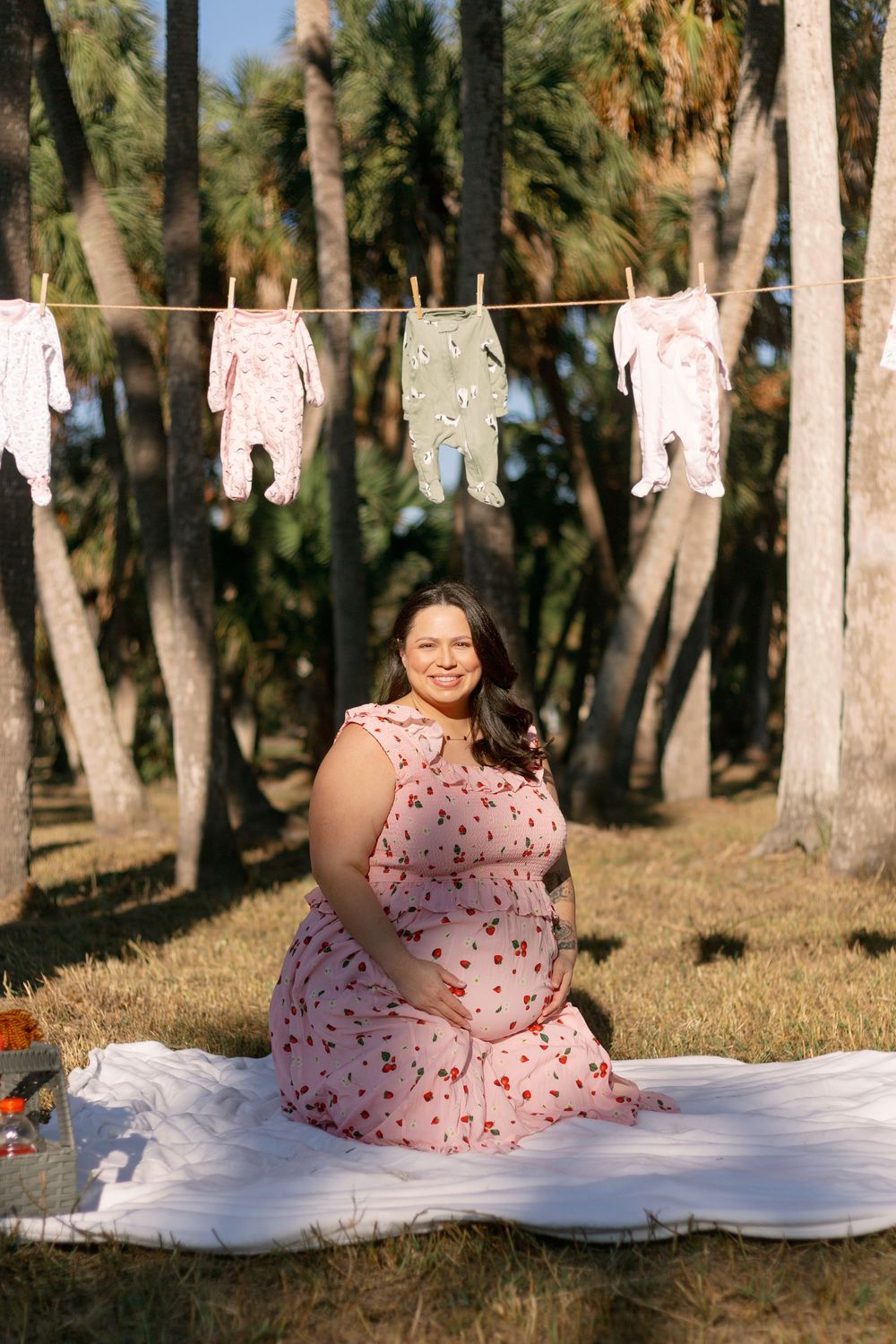 Baby clothes hanging on a clothesline in front of palm trees with someone sitting on a white blanket below.