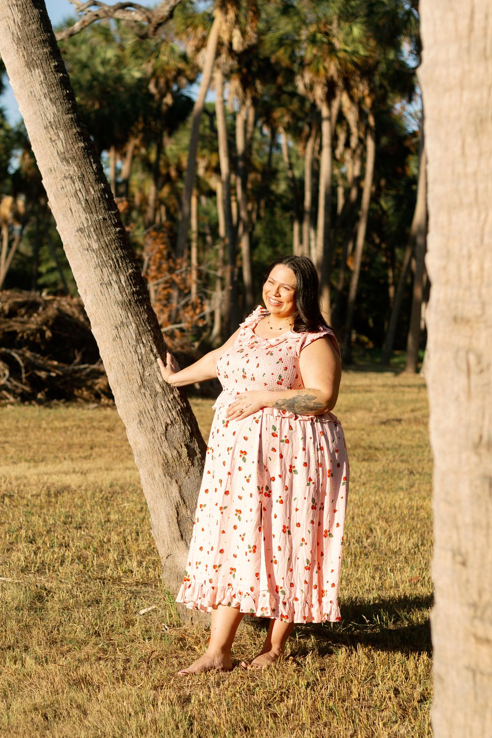 A woman in a pink floral dress stands barefoot next to a palm tree in dappled sunlight.