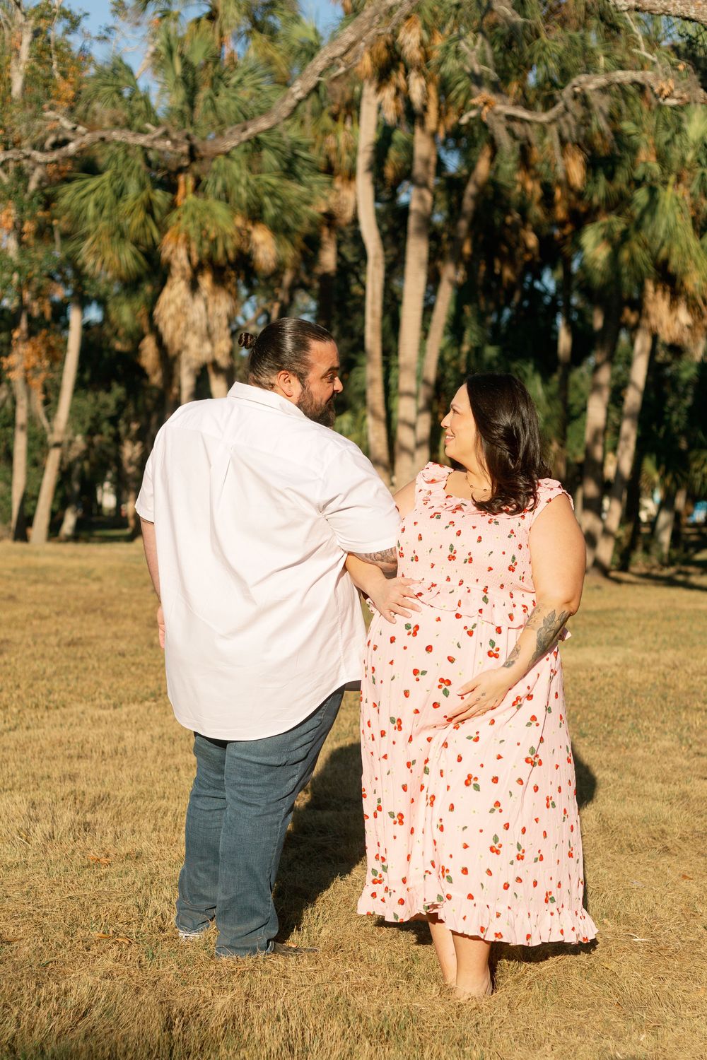 A couple stands together in a grassy field with tall pine trees behind them on a sunny afternoon.