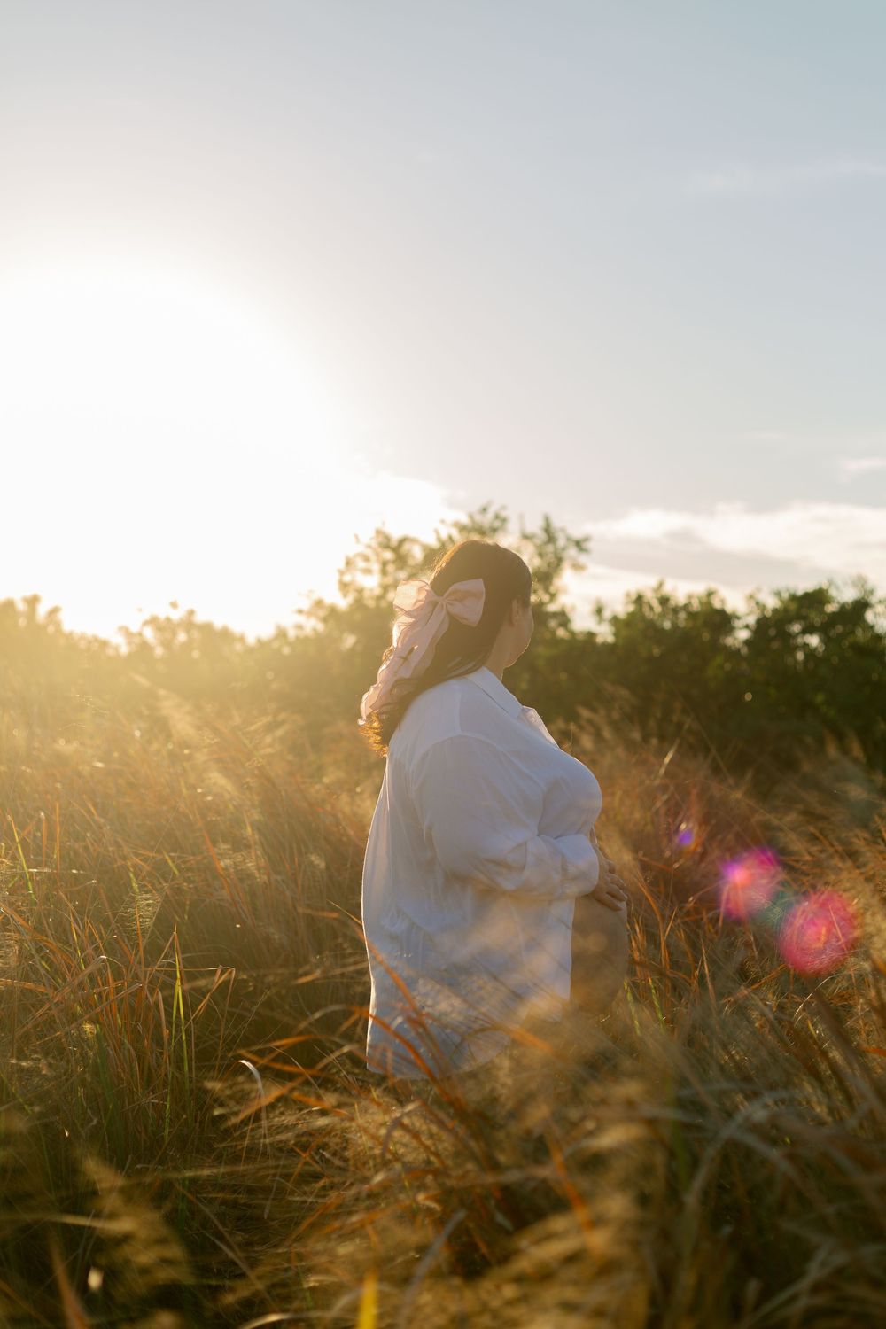 Someone in a white shirt sits in tall grass during golden hour sunset with sun flare and trees in background.