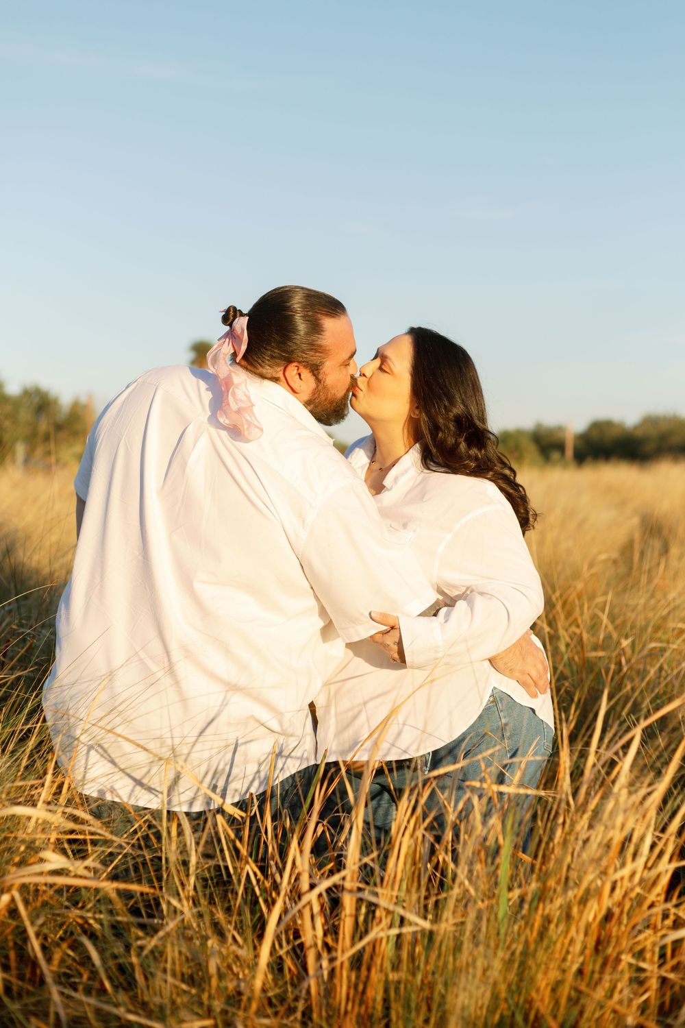 A couple in white outfits sharing a romantic kiss while sitting in a golden sunlit field of tall grass.