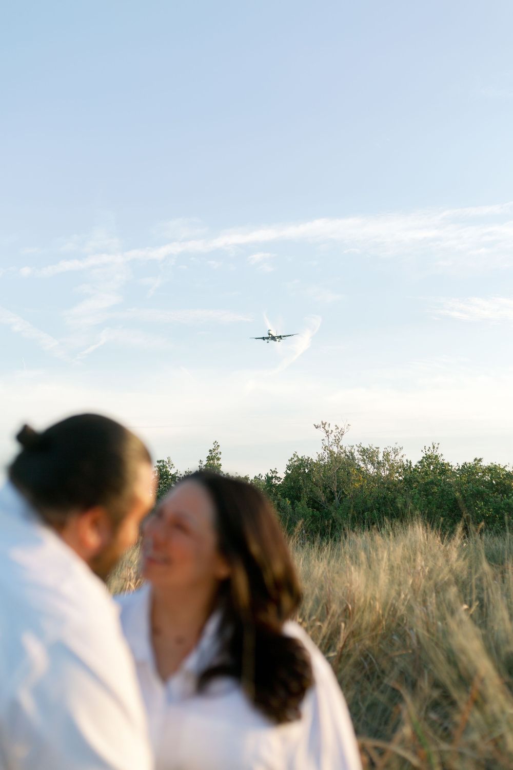 A couple sharing an intimate moment in a grassy field while an airplane soars overhead in the blue sky.