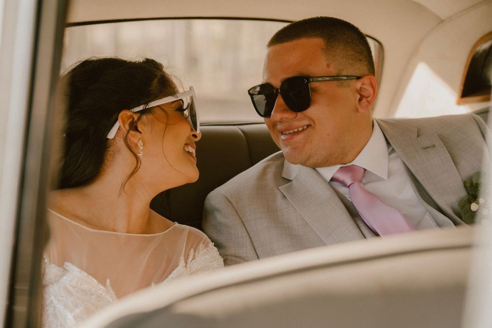 Newlyweds sharing a happy moment while sitting in their wedding car wearing sunglasses.