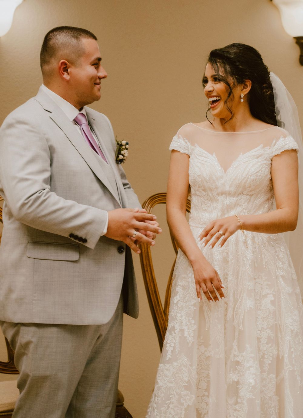 A groom in a light gray suit and bride in a delicate lace dress share a joyful moment during their wedding ceremony.