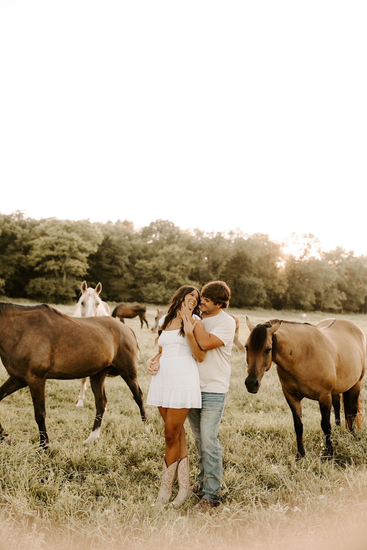 Couple standing among group of horses in sunlit meadow during engagement photos in Gallatin, TN.