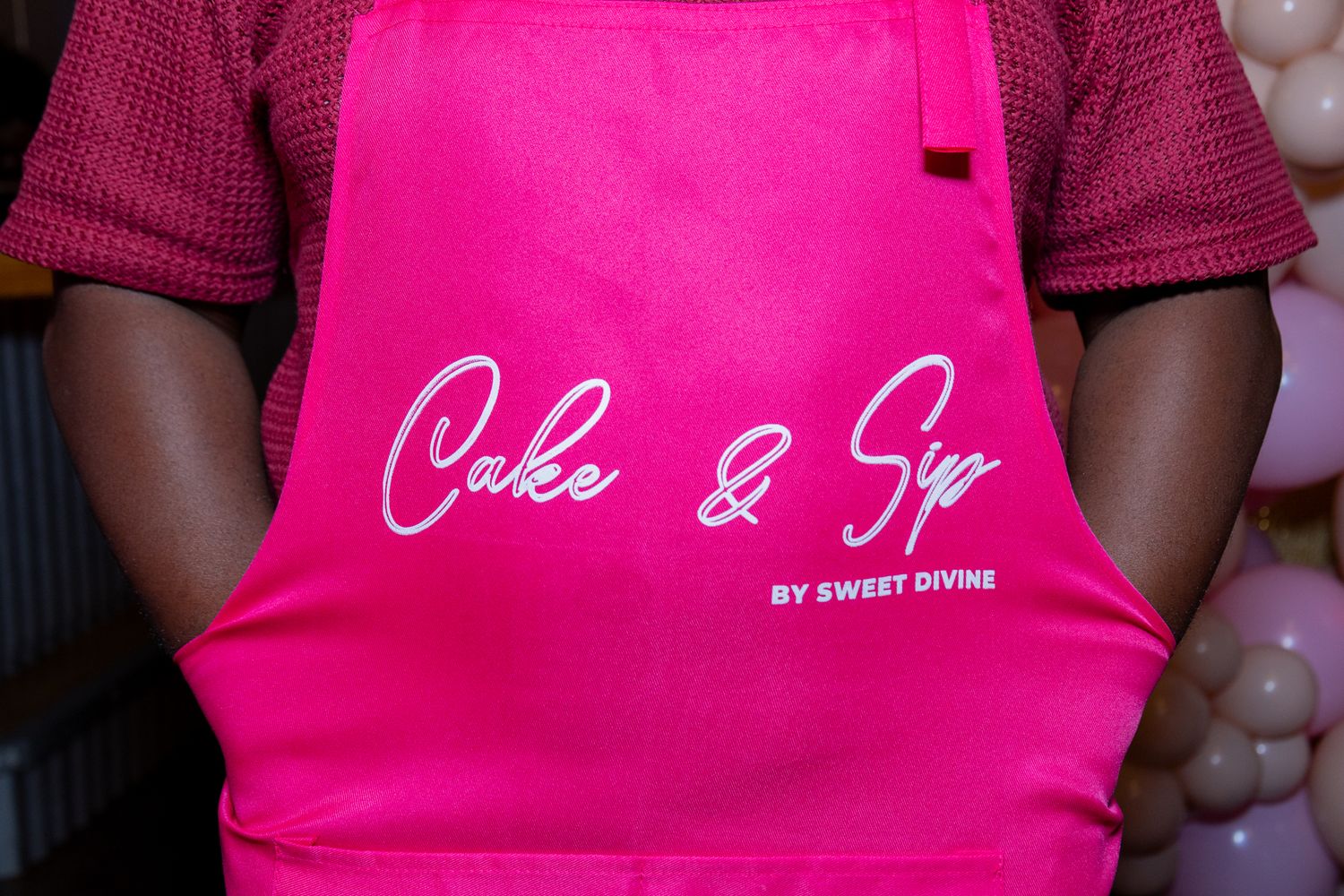 A person wearing a bright pink apron with white script lettering that says Cake & Sip by Sweet Divine.