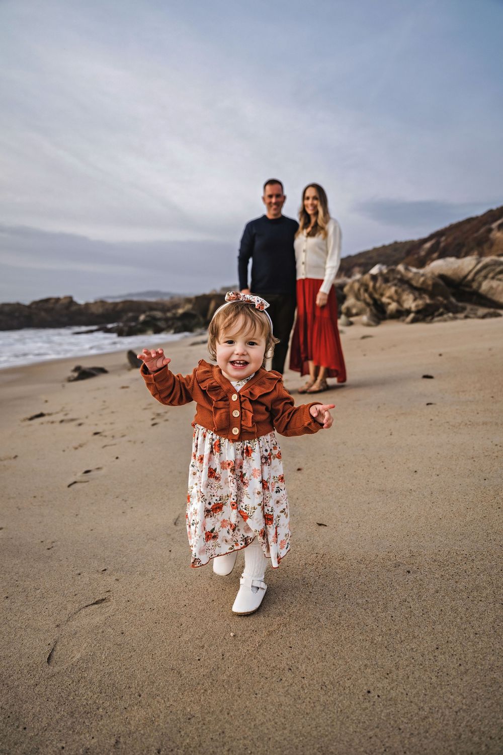 Child in floral dress running joyfully on sandy beach at sunset.