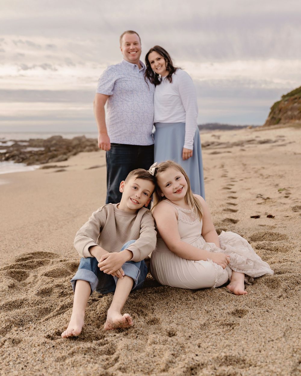 A family sits together on a sandy beach at sunset wearing coordinated light colored outfits.