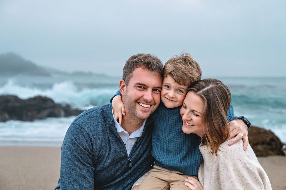 A couple embraces and smiles while spending time together at a scenic beach with waves and rocks in the background.