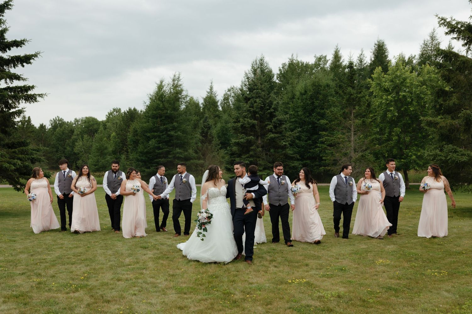 A wedding party stands in formation against a backdrop of tall evergreen trees in a lush green field.