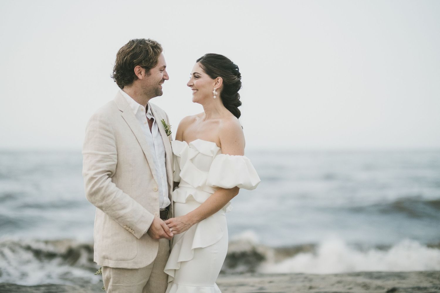 A couple embraces each other on a rocky beach with ocean waves in the sunset background.