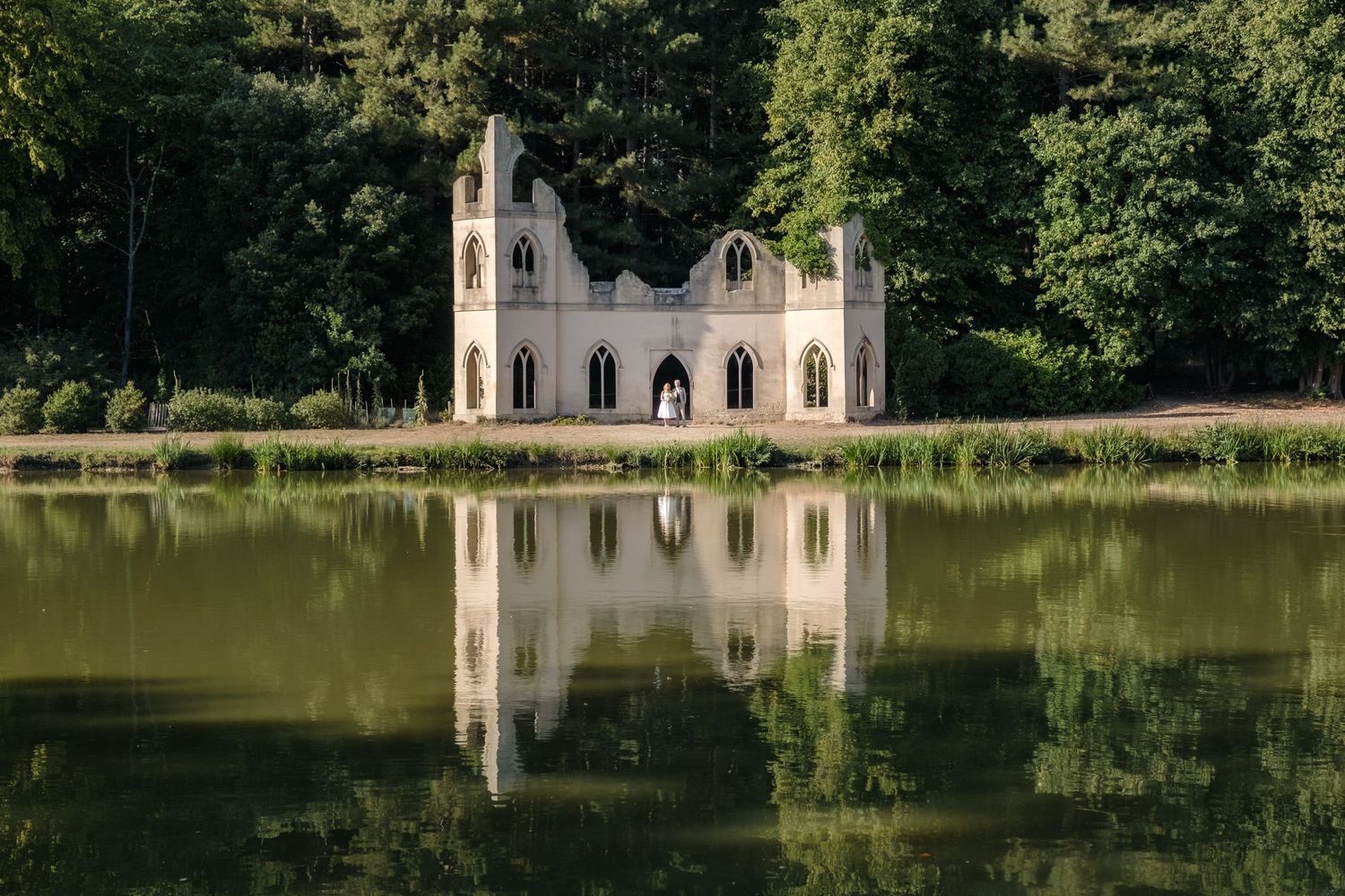 A serene white chapel with arched windows reflects on the calm waters of a lake surrounded by lush green trees.