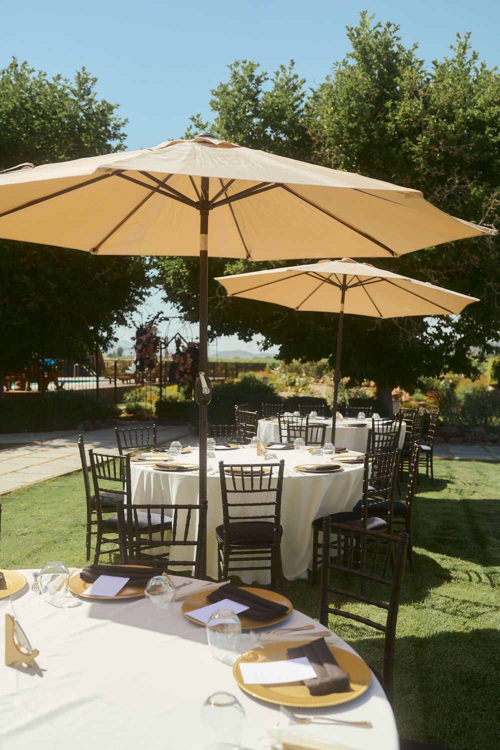 Outdoor wedding reception tables with white linens, gold charger plates, and beige patio umbrellas in a garden setting.