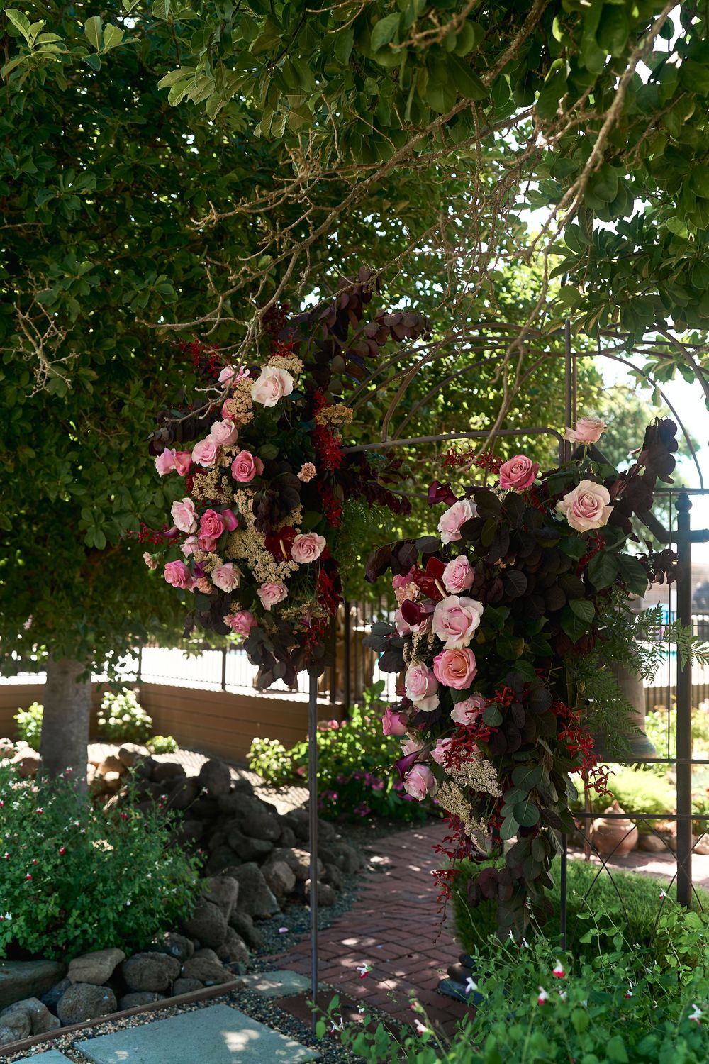 Beautiful floral wedding arch adorned with pink and burgundy roses surrounded by lush green garden foliage.