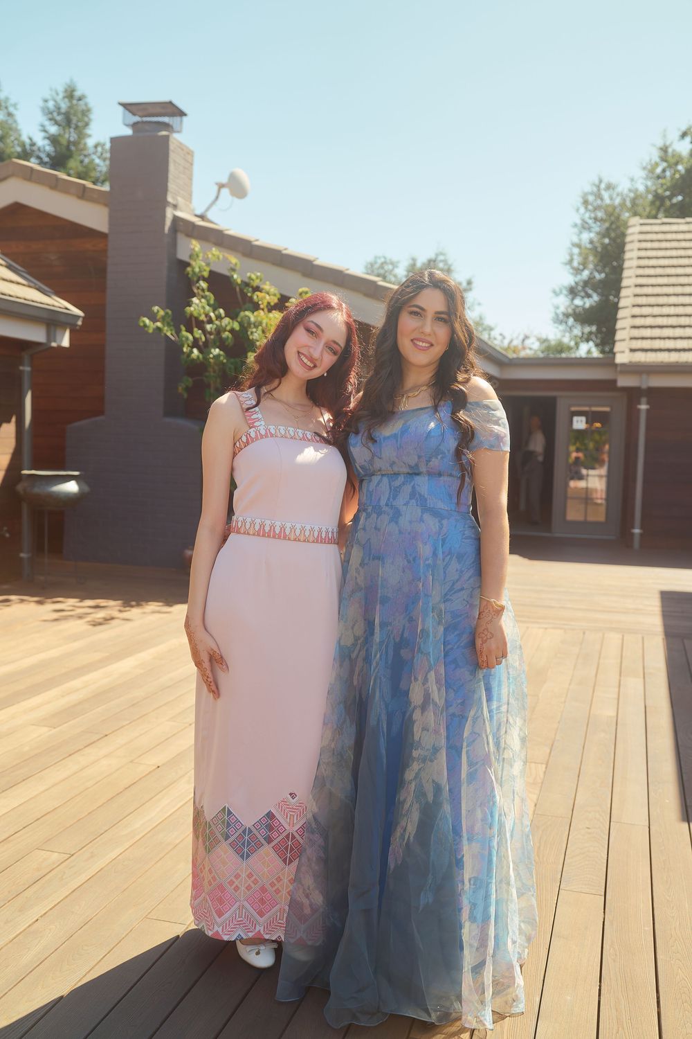 Two young women in elegant formal gowns, one pink and one blue floral, posing together on a sunny wooden deck.