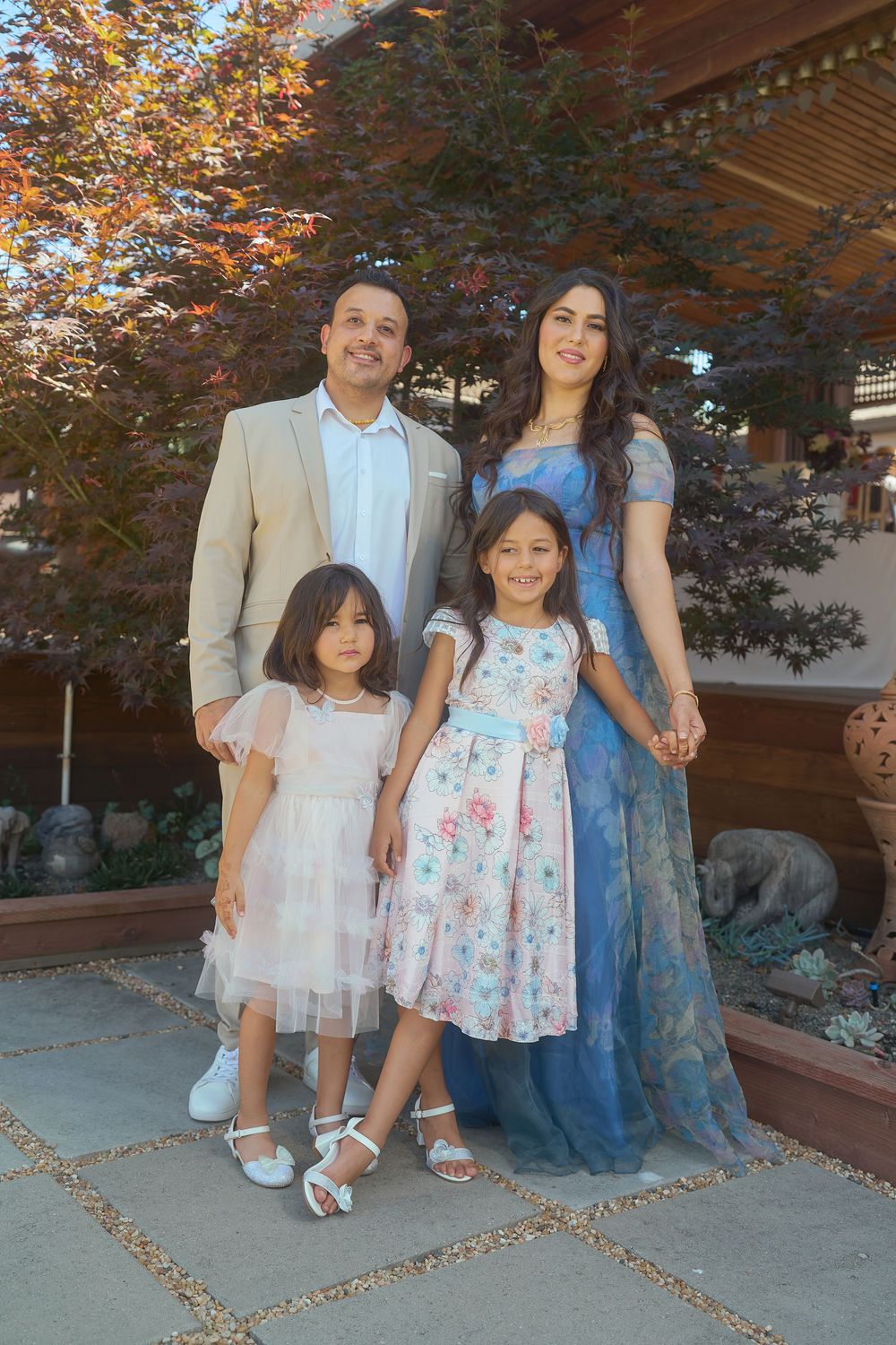 Family of four posing outdoors at a wedding, parents in formal attire with two young daughters in floral dresses.
