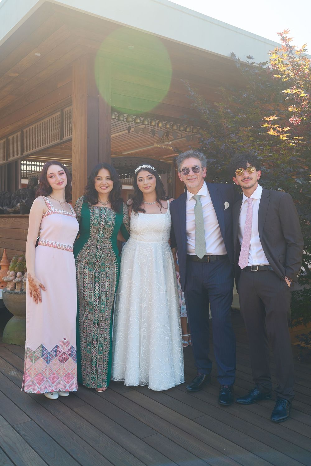 Wedding party group of five posing outdoors, including bride in white gown and guests in formal attire.