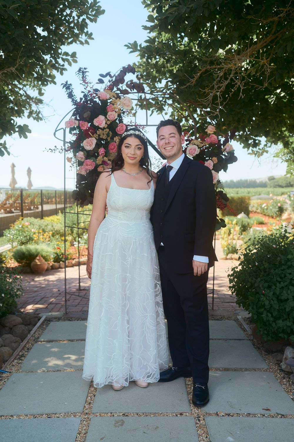 Bride and groom posing together under a floral arch with pink roses in a lush garden setting.