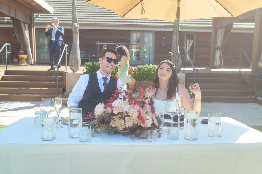 Bride and groom smile at floral-decorated sweetheart table at sunny outdoor wedding.
