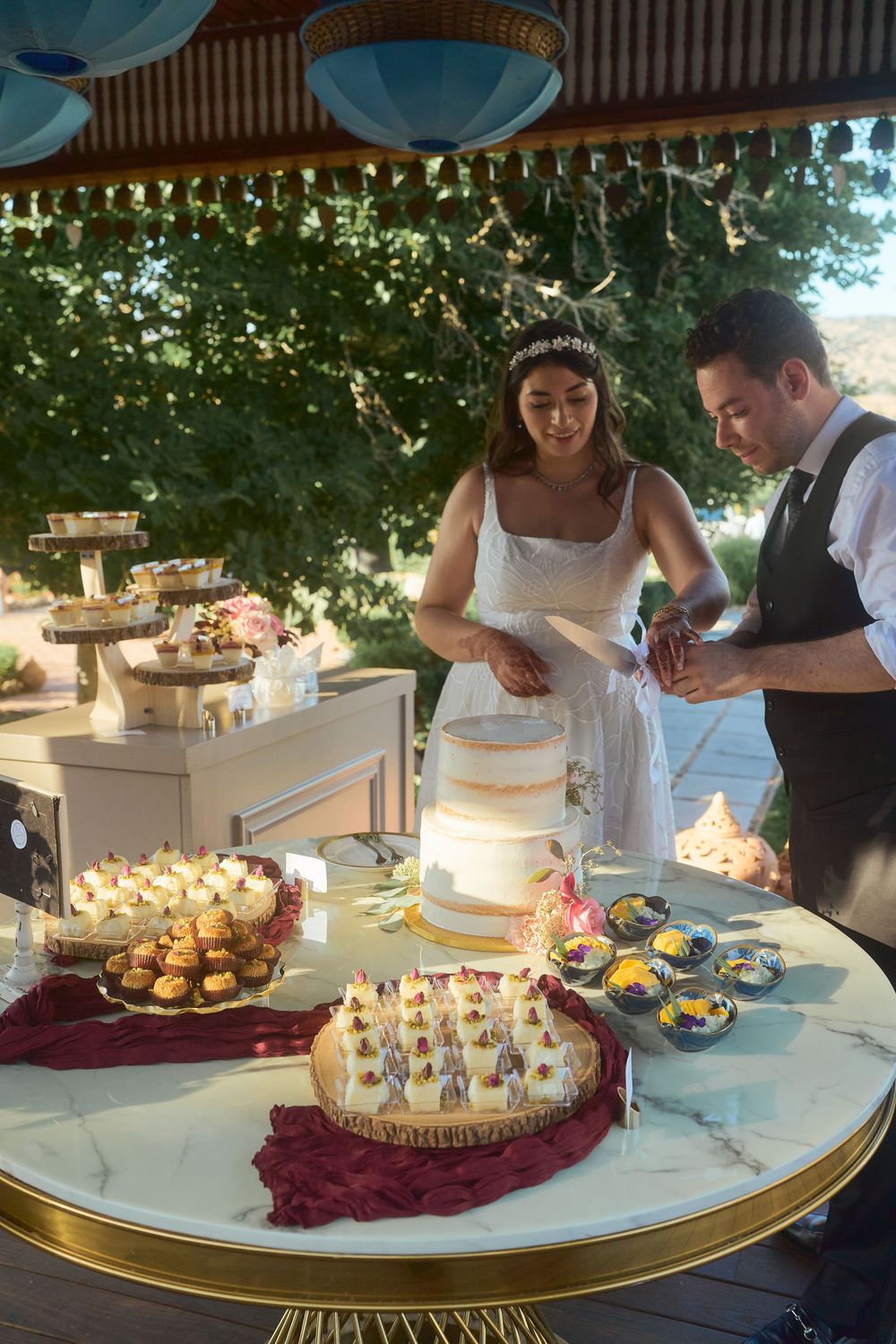 Bride and groom cutting their wedding cake at an elegant outdoor dessert table filled with cupcakes and treats.