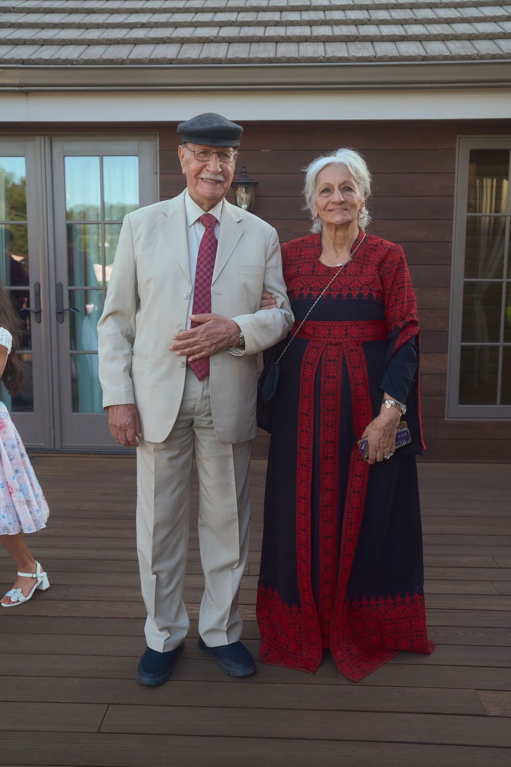 Elegantly dressed elderly couple posing together on a wooden deck at an outdoor wedding celebration.